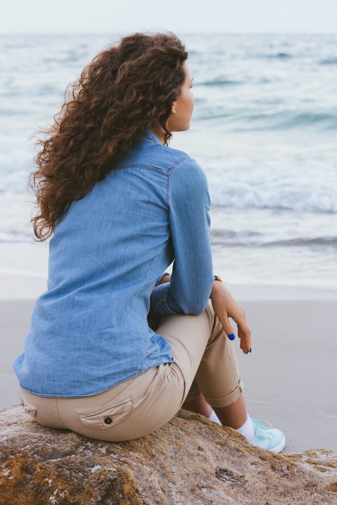 Beautiful slim woman with curly hair sitting on a rock by the sea. She is dressed in beige trousers and a denim shirt. Back view.