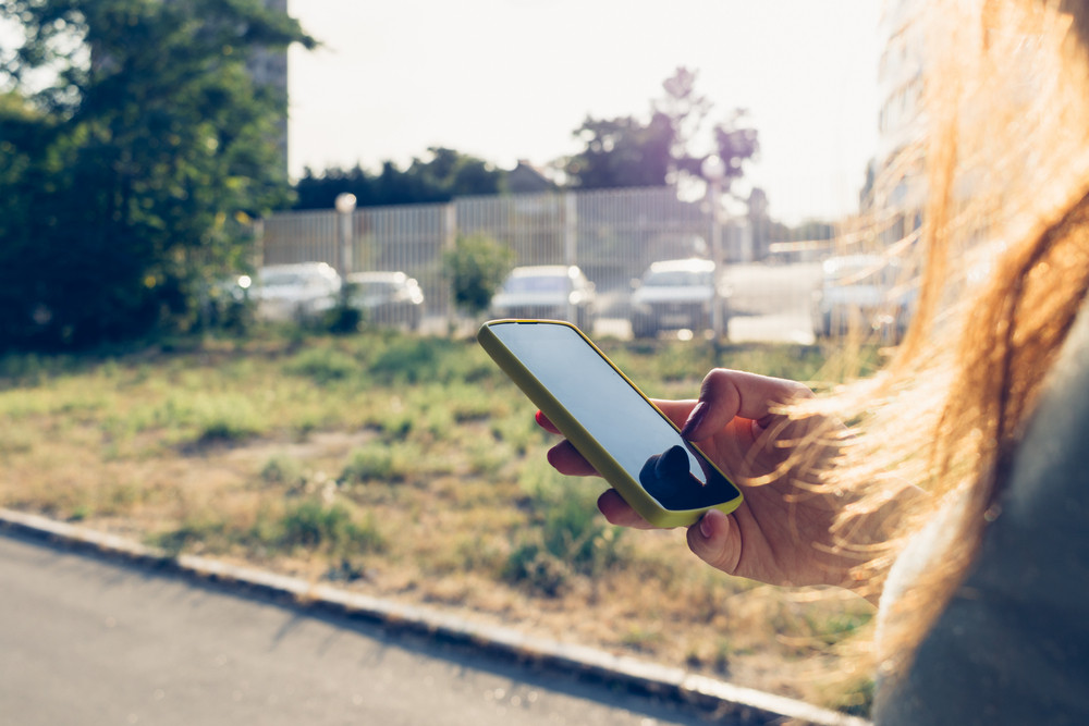 Close-up of a mobile phone in a female hand on the background of the city in the sunlight. Woman walks down the street and looking at the phone\'s screen.