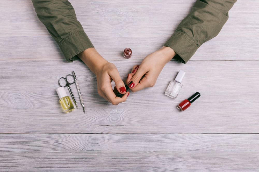 Female hands make manicure and paint nails with red lacquer. On the table are bottles of nail polish, scissors and other tools, top view.