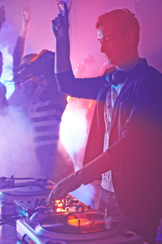 Male deejay mixing sounds on turntables with group of people dancing on background
