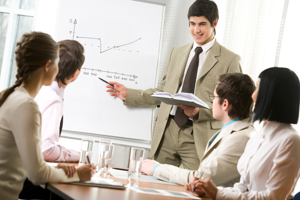 Portrait of handsome businessman pointing at graph on board and looking at his co-workers with smile