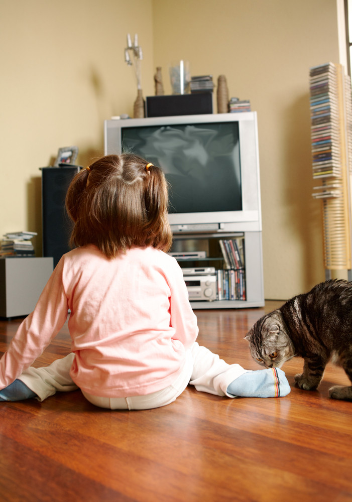 Rear view of little girl sitting on the floor and watching TV with cat near by