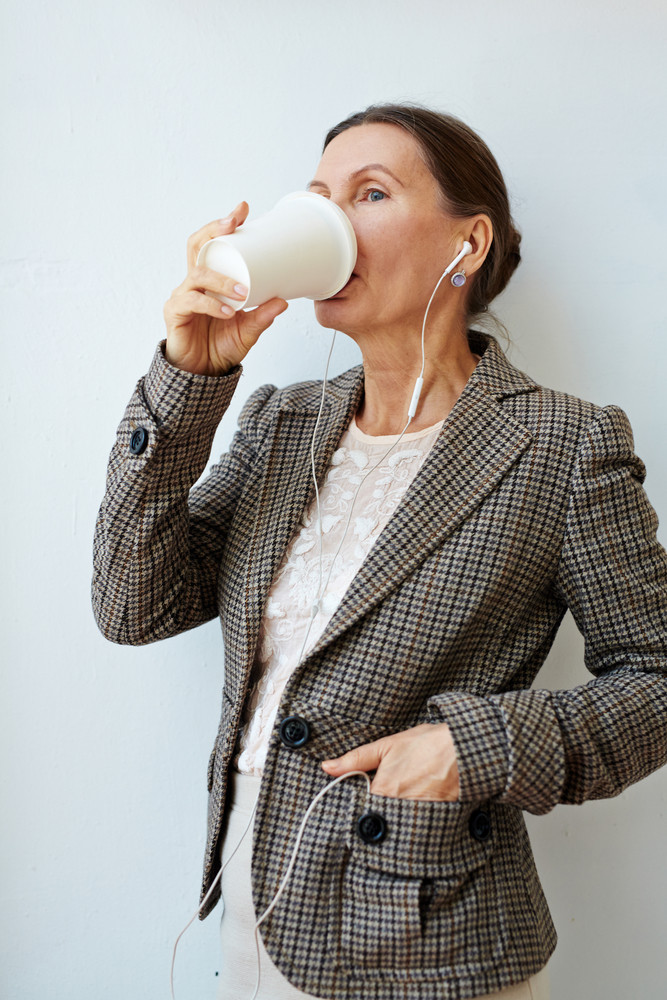 Beautiful mature entrepreneur standing against white wall with headphones in ears, drinking delicious coffee from paper cup and looking away, portrait shot