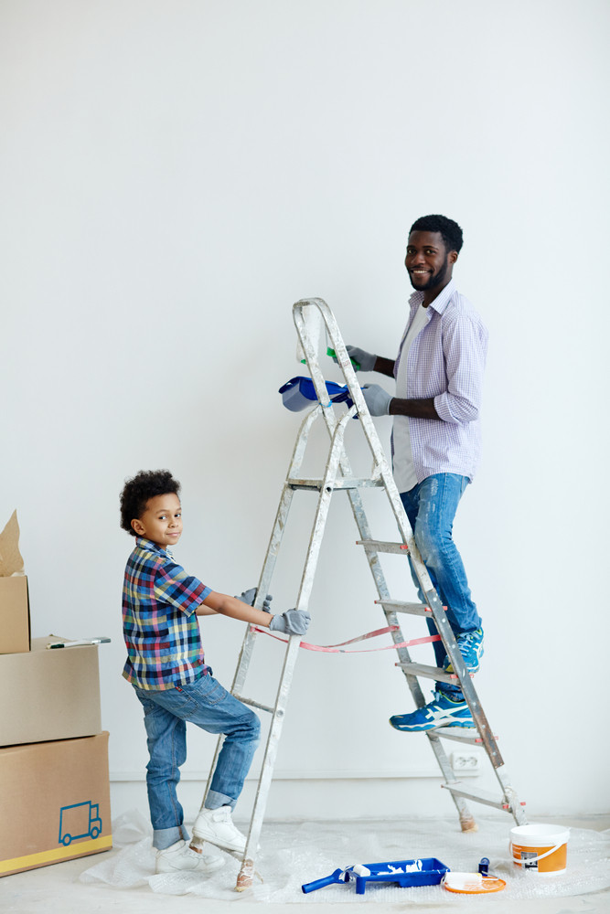 Boy holding ladder while his father painting wall in new flat