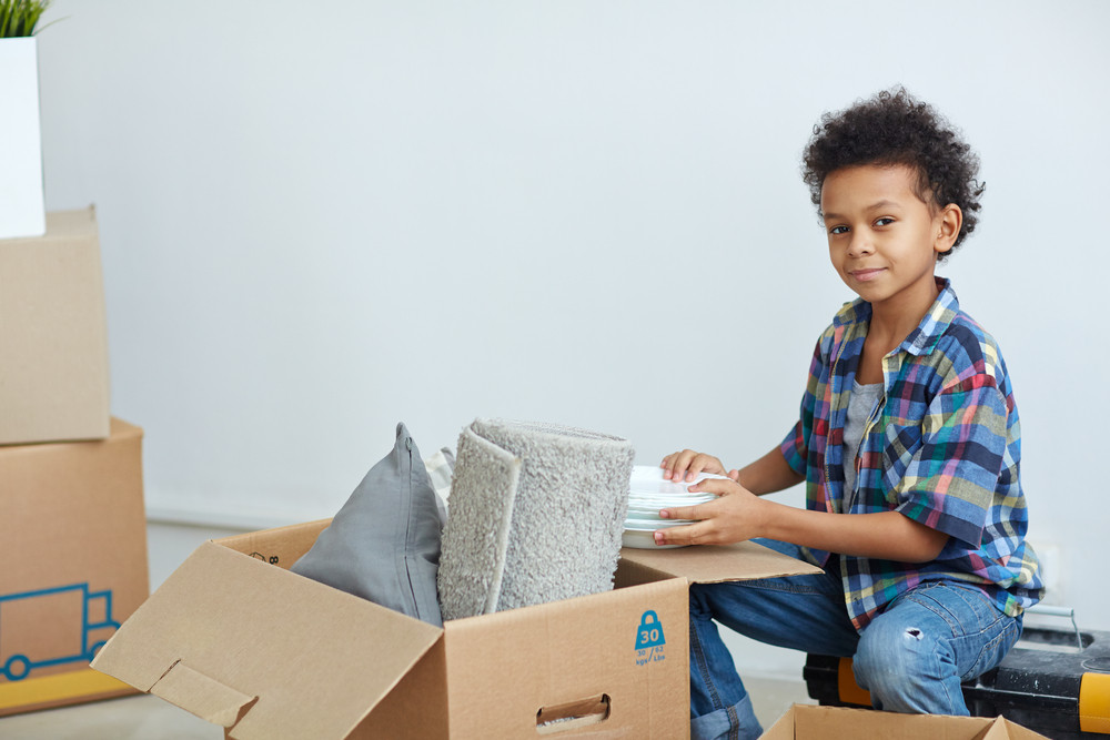 Boy with stack of plates sitting by open box with grey rolled rug and pillow