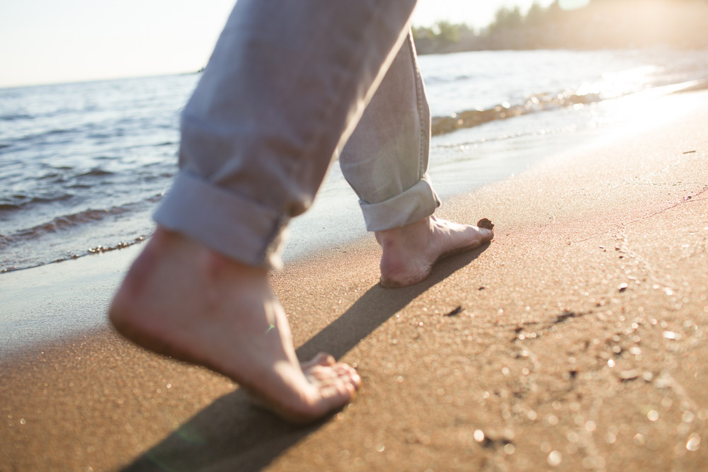 Feet of man walking down wet sand along coastline in the morning