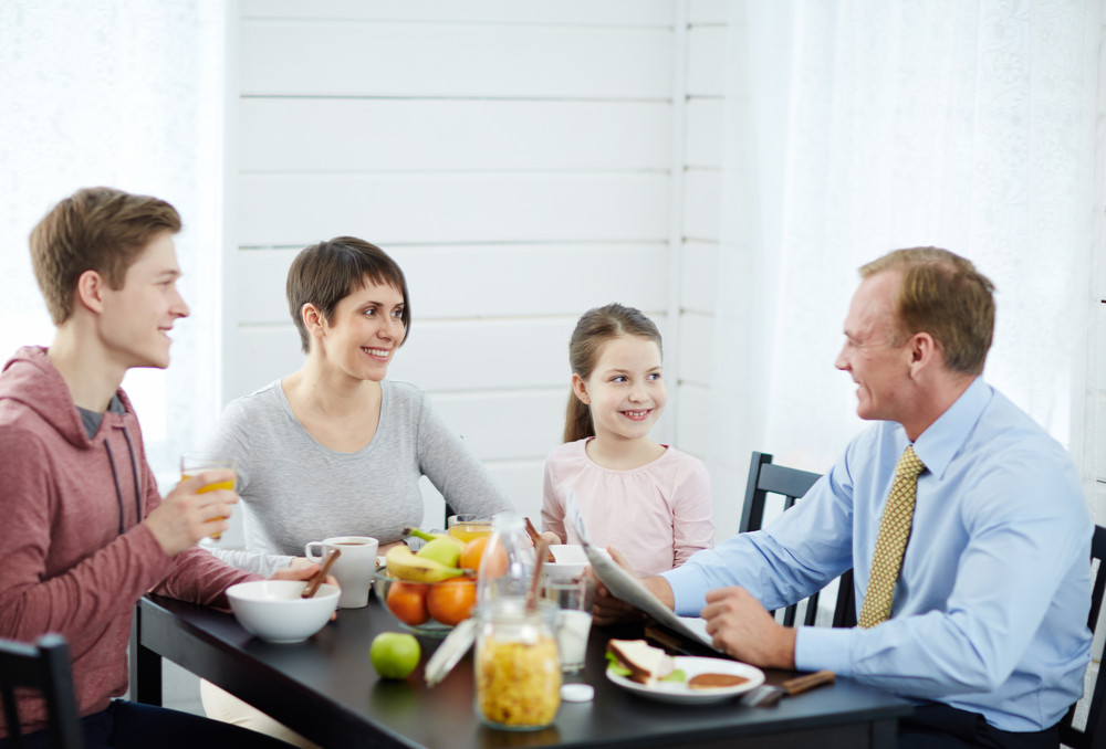 Lovely family moments at breakfast time: pretty middle-aged mother, little daughter and teenage son looking at head of family with wide smiles while he entertaining them with funny story