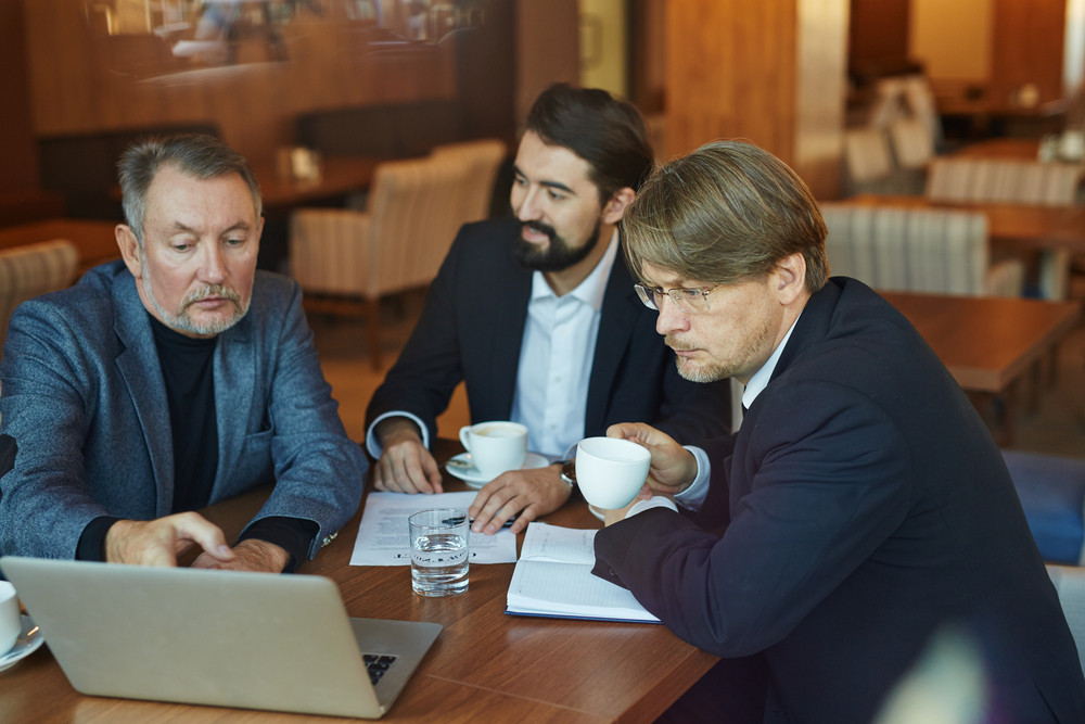 Three hard-working bearded businessmen gathered together in cozy cafe and calculating profit margins from their startup launching, waist-up portrait