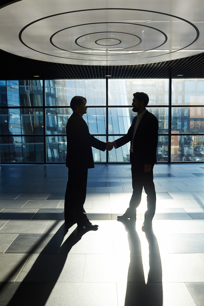 Two middle-aged businessmen standing against panoramic window of office lobby and shaking hands, full-length portrait