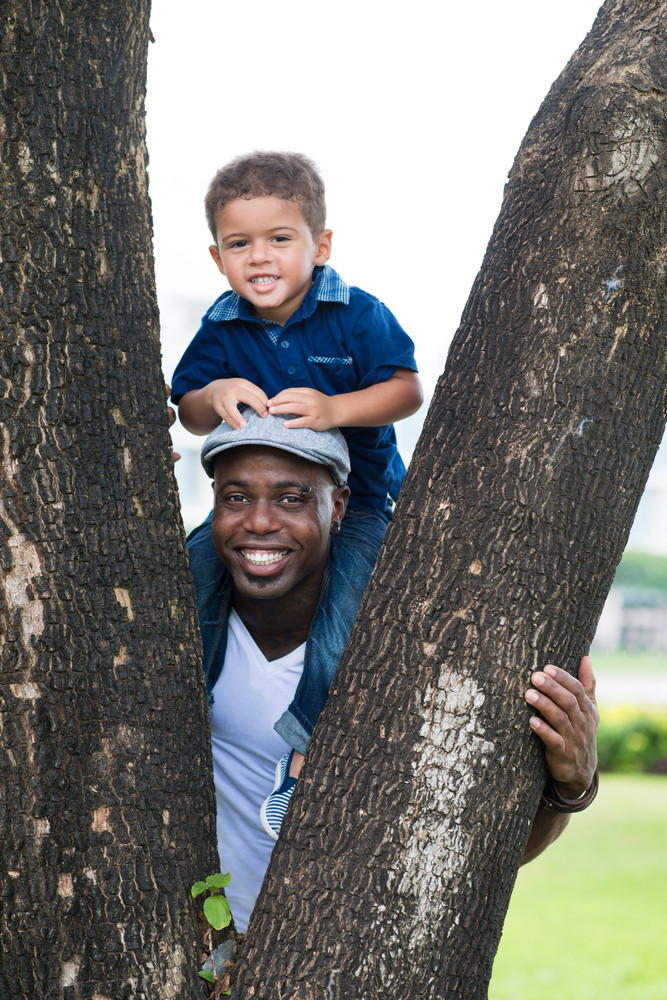Vertical portrait of a black father carrying his son on his shoulders standing behind the tree