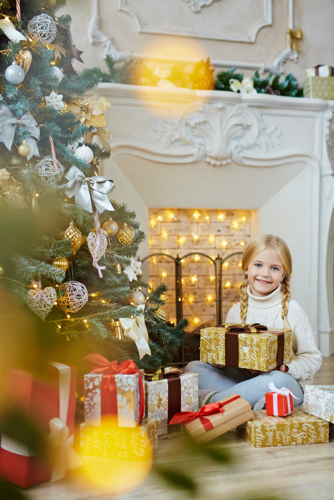 Young girl with gift-box sitting by fireplace and decorated Christmas tree