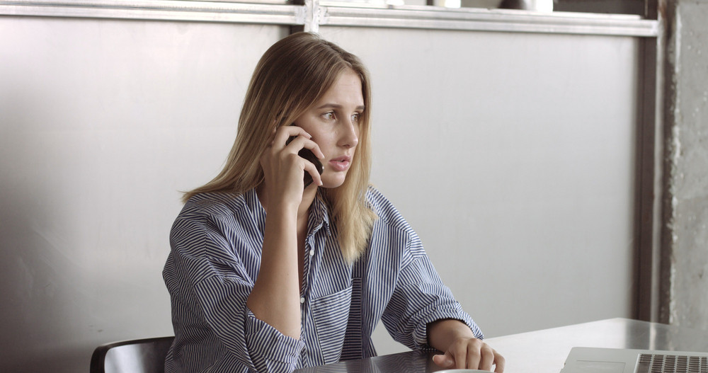 Blond model in a relaxed button-up shirt switches between her laptop and her phone at work