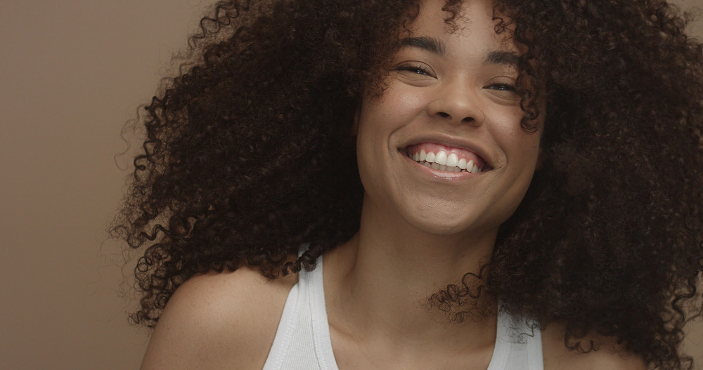 mixed race black woman portrait with big afro hair, curly hair in beige background smiling and laughing closeup
