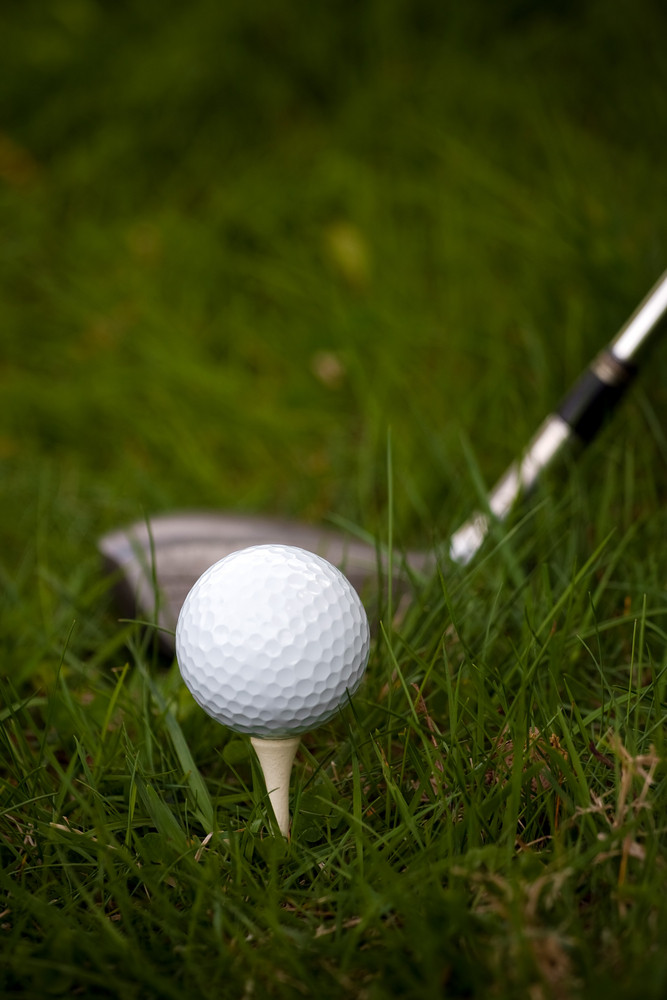 A white golf ball set up on the tee in the rough with a driver about to swing. Shallow depth of field.
