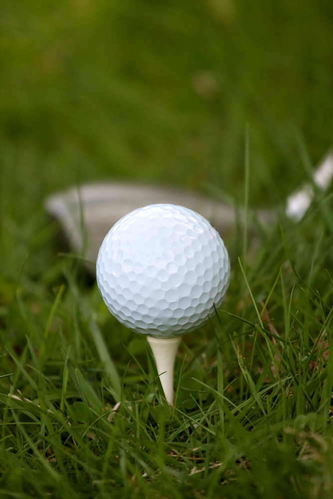 A white golf ball set up on the tee with a driver about to swing. Shallow depth of field.