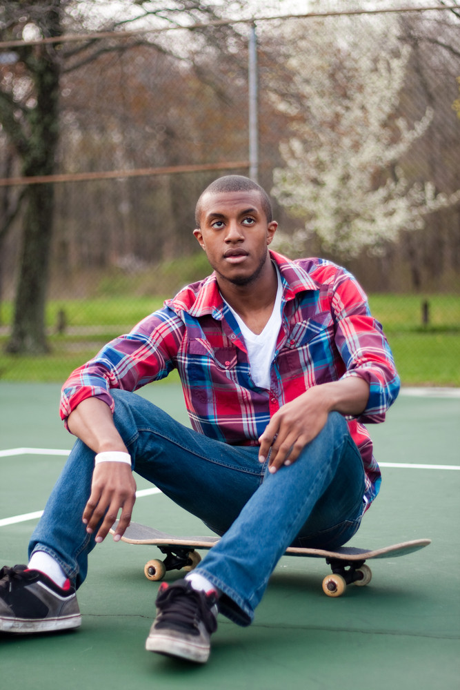 A young man hanging out in the tennis courts sitting on his skateboard.