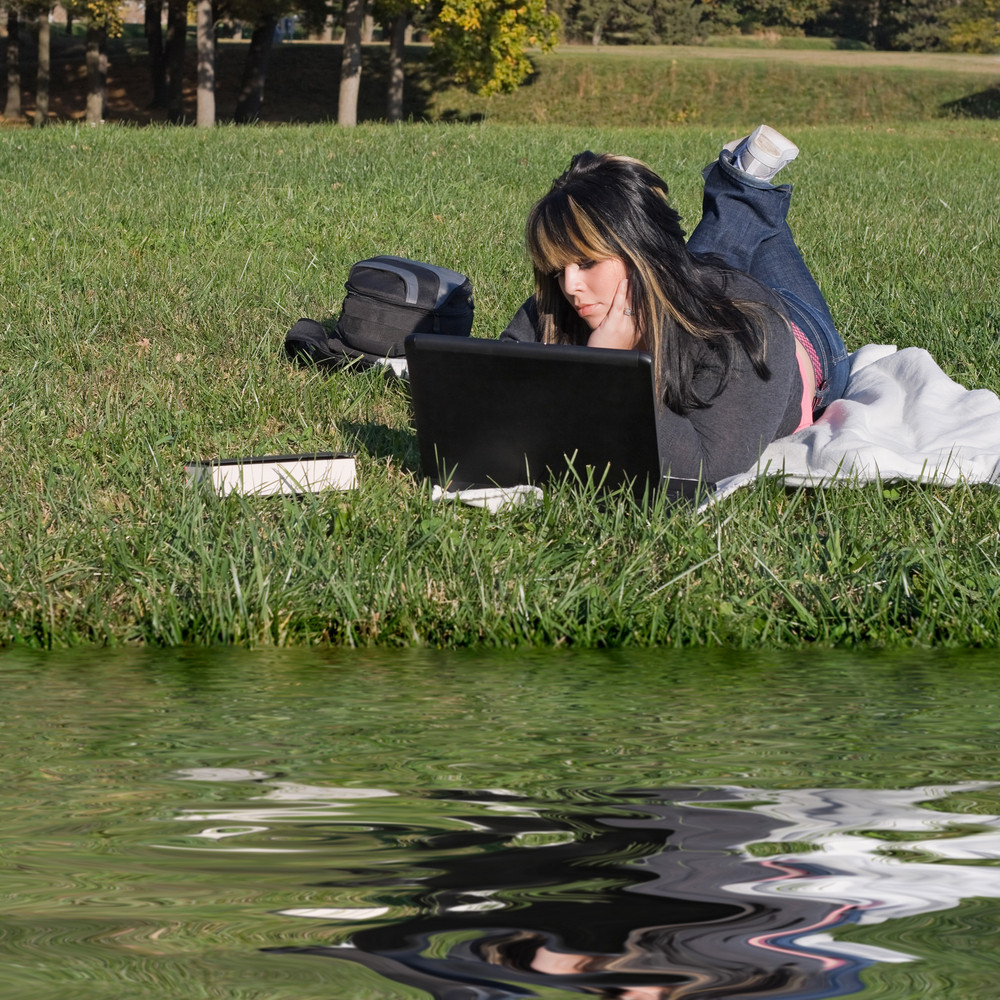 A young student using her laptop computer while laying in the grass on ...