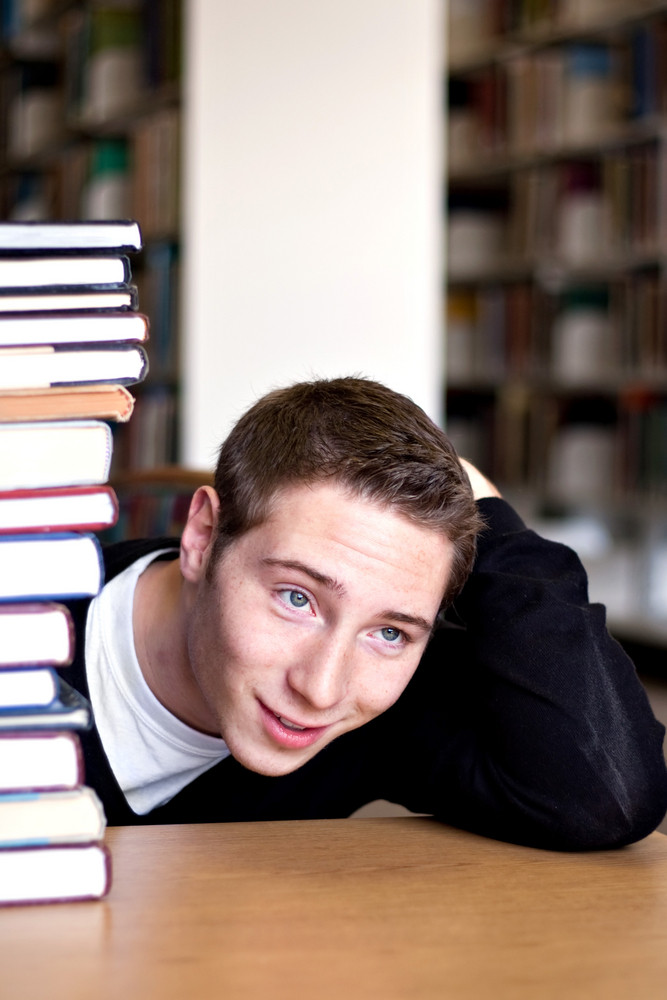 An overwhelmed student with a high pile of textbooks he has to go through to do his homework.