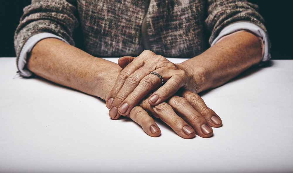 Close-up studio shot of a senior woman\'s hands resting on grey surface. Old lady sitting with her hands clasped on a table.