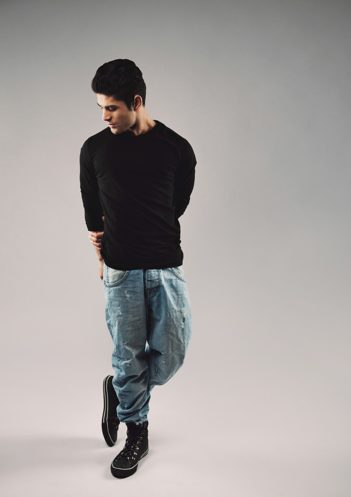 Full length portrait of hispanic young man in casuals posing. Male model looking down while standing on grey background.