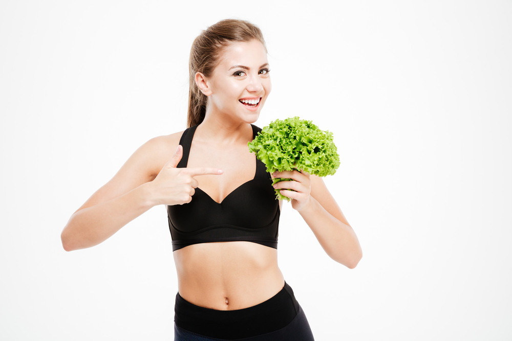 Attractive smiling fitness woman pointing finger at lettuce leaves isolated on a white background