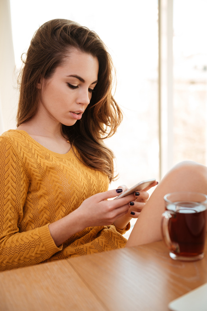 Attractive young woman reading message on her cellphone and drinking tea indoors