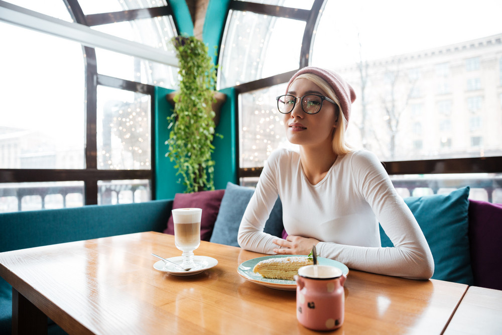 Beautiful young woman in hat and glasses drinking coffee and eating cake in cafe