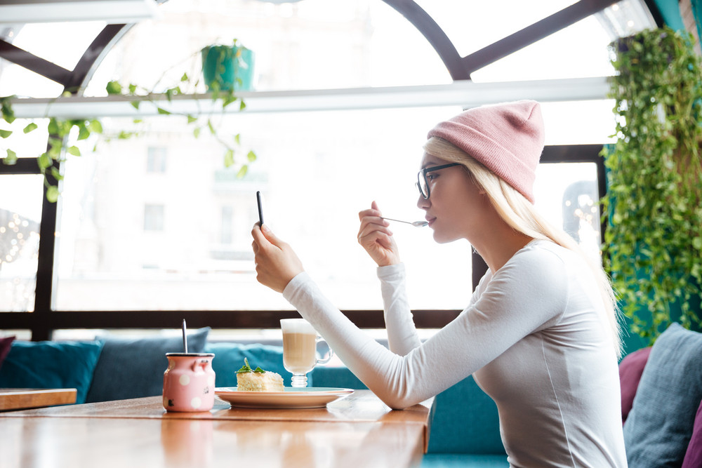 Beautiful young woman in hat and glasses eating dessert and using cell phone in cafe