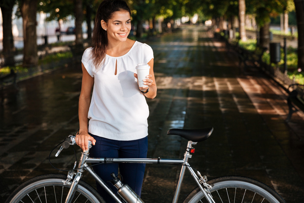 Cheerful brunette woman with takeaway coffee cup and bicycle in the park