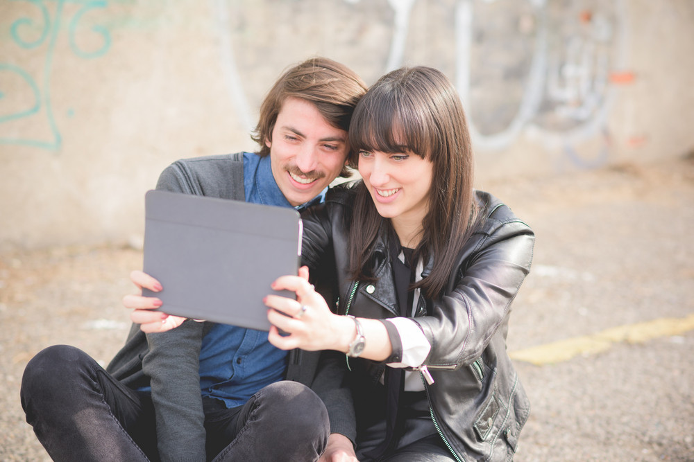 Couple of young beautiful caucasian man and woman seated on a sidewalk in the city using a tablet taking a selfie - social network, technology, communication concept