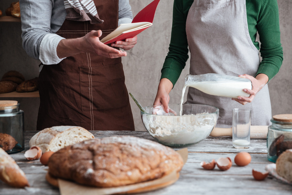Cropped image of loving couple bakers standing near bread and cooking. Man holding notebook.