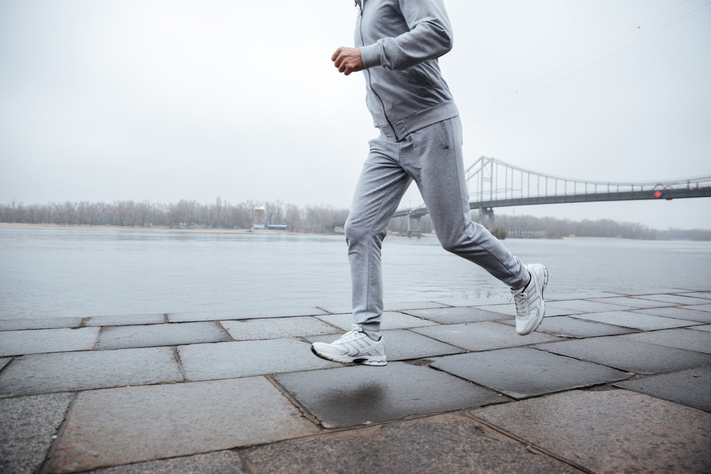 Cropped image of man in gray sportswear running near the water. Side view