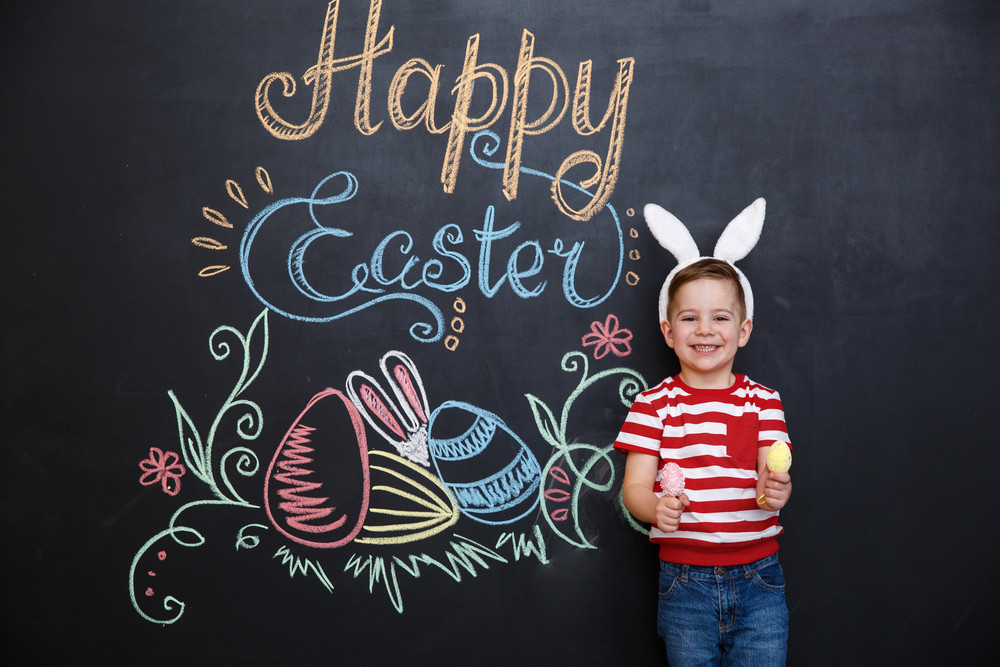 Cute little boy wearing rabbit ears and holding two eggs while standing inside big cracked eggshell over chalk board with easter doodles background