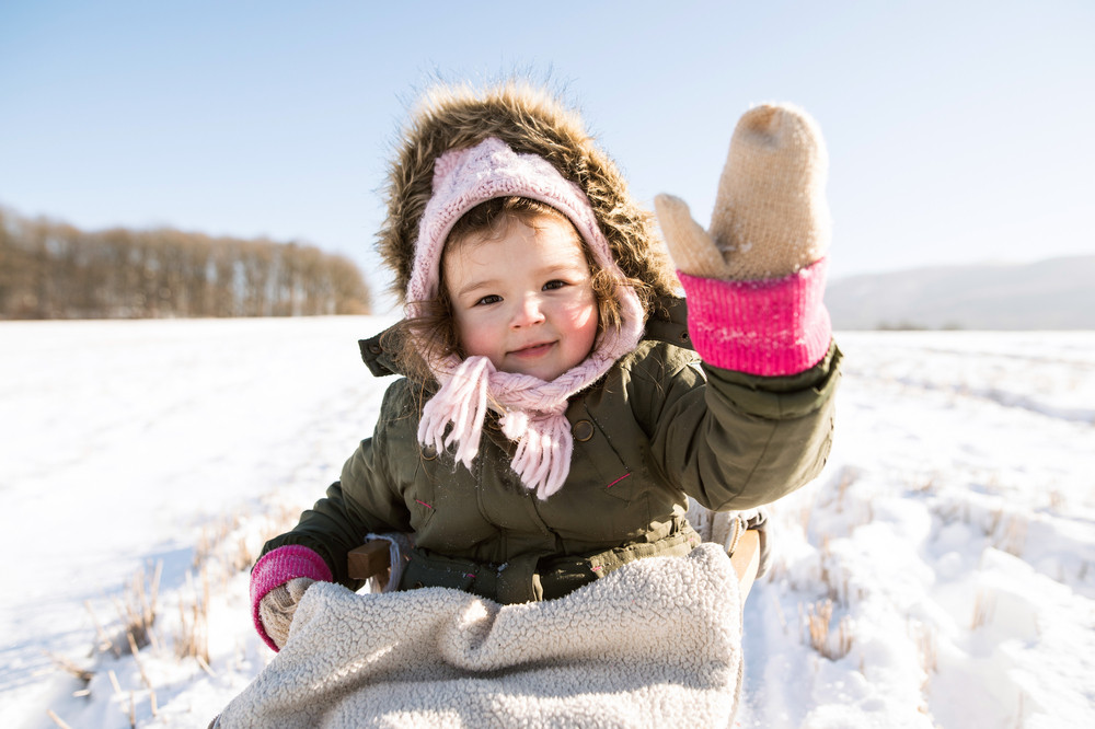 Cute little girl outside in winter nature on sunny day, sitting on sledge, waving