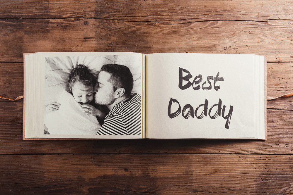 Fathers day concept. Photo album, black-and-white picture of daughter lying in bed, her father kissing her on cheek. Studio shot on wooden background.