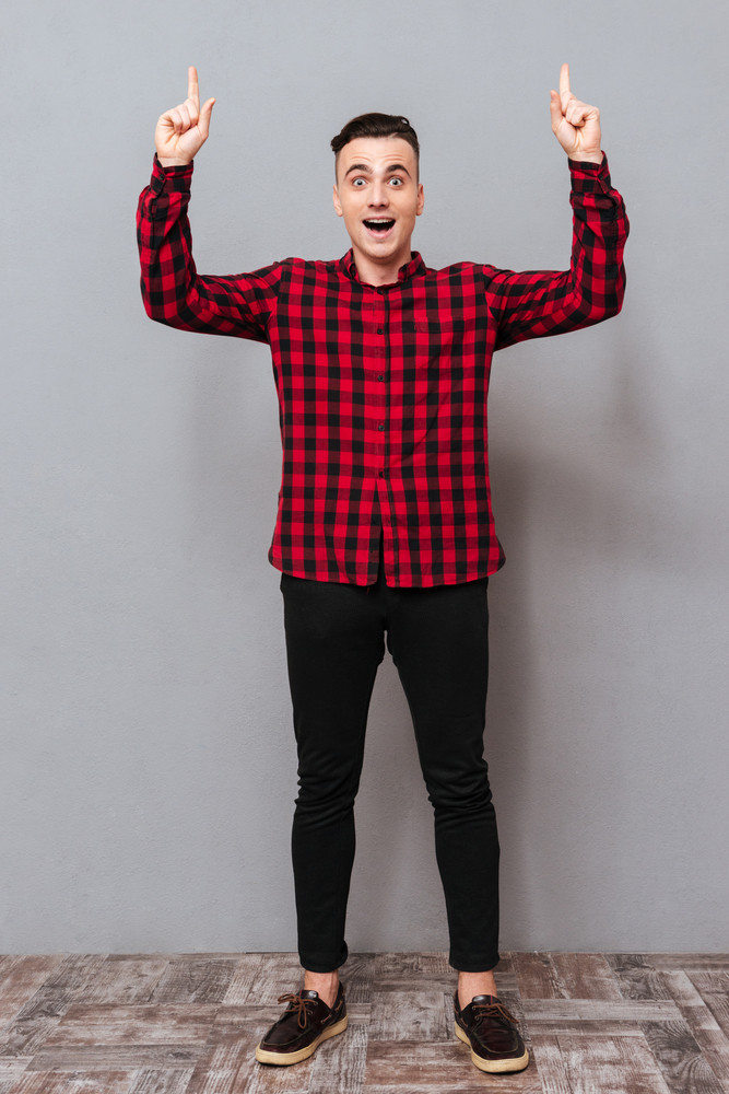 Full length surprised young man in shirt and black jeans pointing up and looking at camera. Isolated gray background