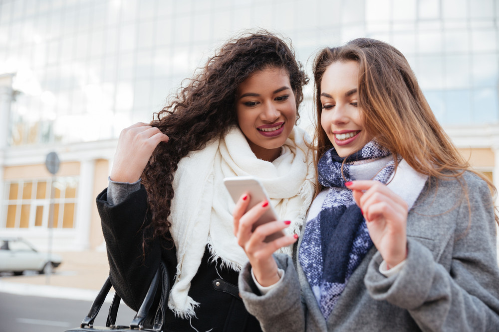 Gorgeous two happy young girlfriends looking on a smartphone. Caucasian girl wearing scarf in a cage print. African lady wearing burgundy scarf.