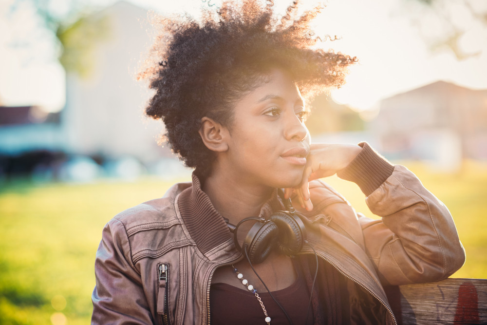Half length of beautiful black curly hair african woman listening music with headphones in city back light - music, technology, serenity concept
