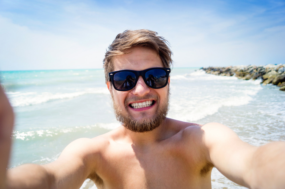 Handsome hipster man on beach, wearing sunglasses, smiling, taking ...