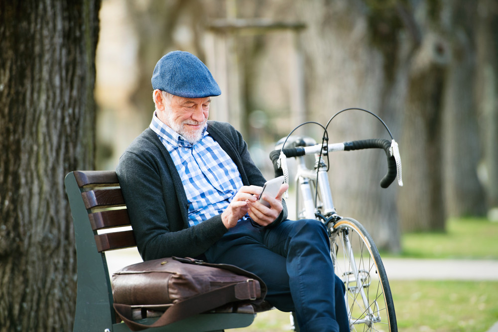 Handsome senior man with bicycle in town park sitting on bench, holding smart phone, texting. Sunny spring day.