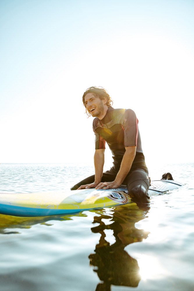 Handsome Smiling Curly Surfer Man In Swimwear Sitting On