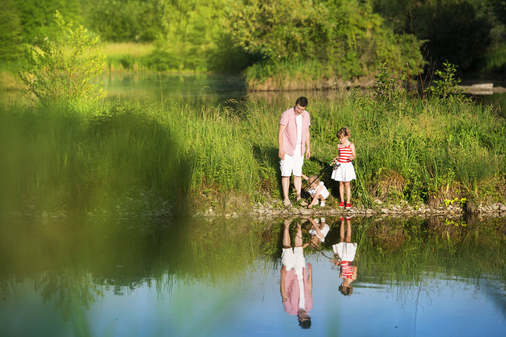 Happy young father fishing on the lake with his little daughters, reflecting in the water
