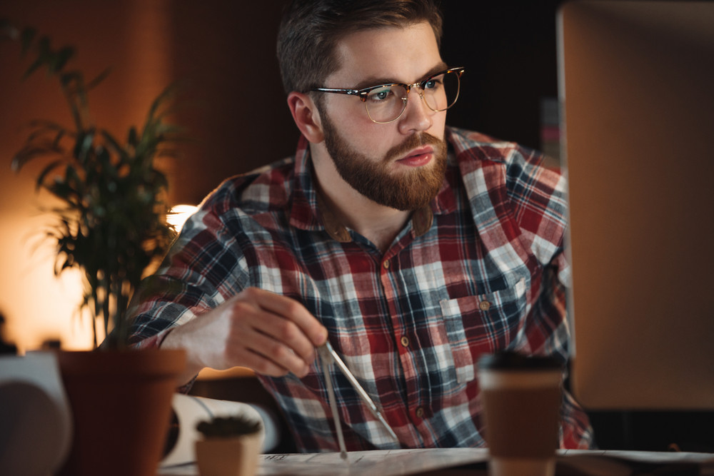 Image of bearded designer dressed in shirt in a cage print and wearing glasses working late at night and looking at computer.