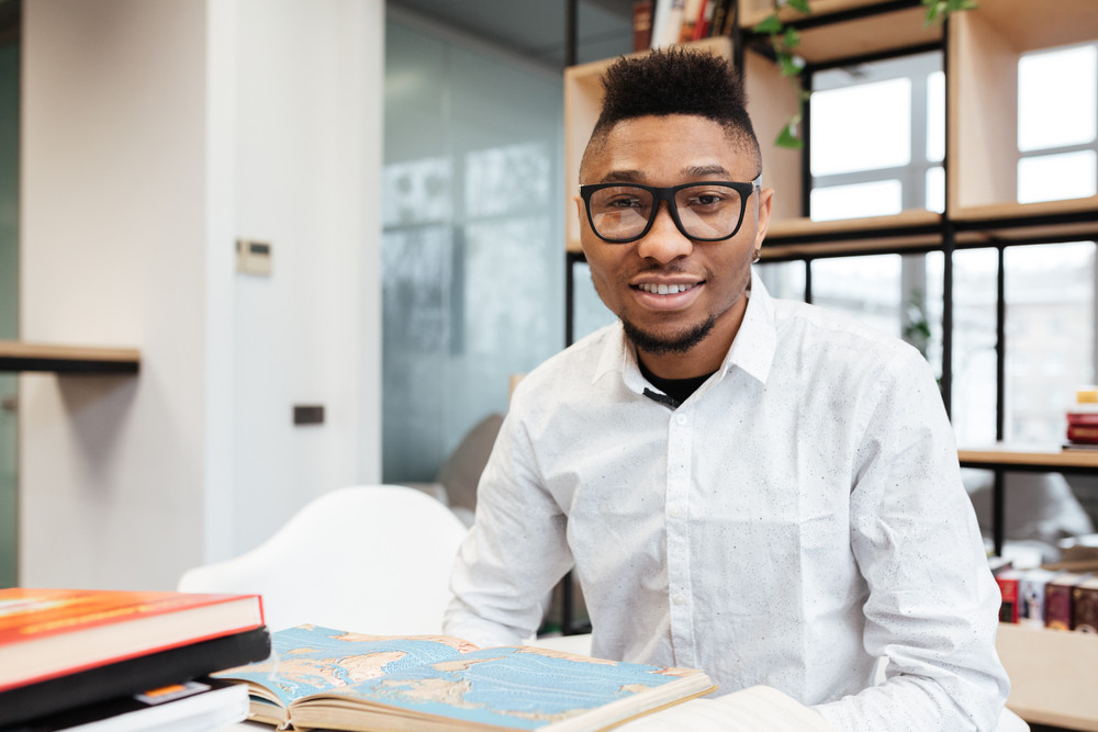 Image of cheerful african student wearing eyeglasses in library learning education material with books. Looking at camera.