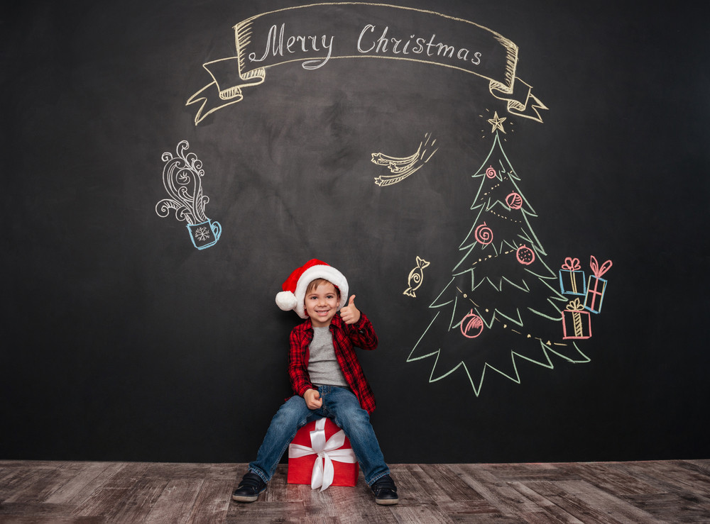 Image of happy child wearing hat sitting on big gift near Christmas tree drawing on blackboard and making thumbs up gesture. Looking at camera.
