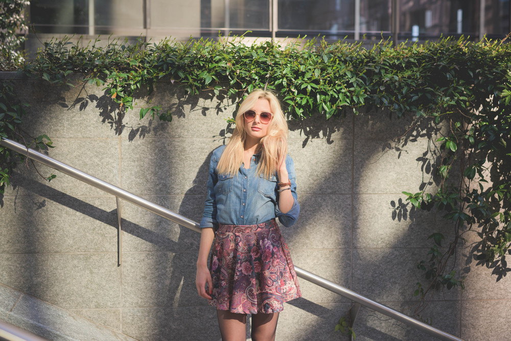 Knee figure of a young beautiful blonde caucasian girl posing in the city wearing a jeans shirt and a floral skirt looking in camera leaning on a wall - youth, freshness concept