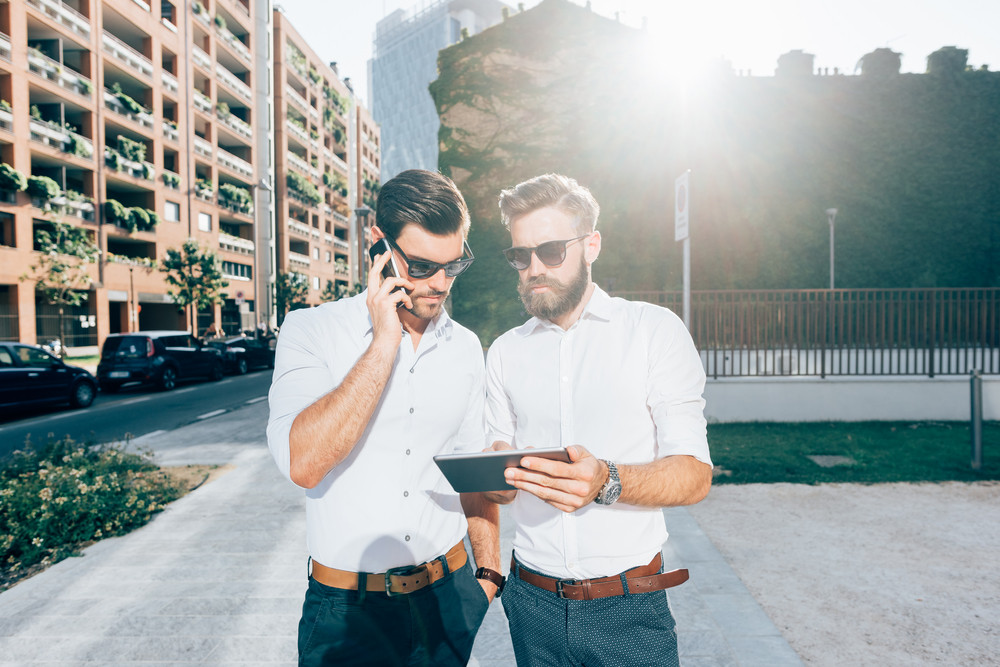 Knee figure of two handsome bearded business man outdoor in the city, one talking smart phone, the other using a tablet hand hold - business, start up, finance, technology concept