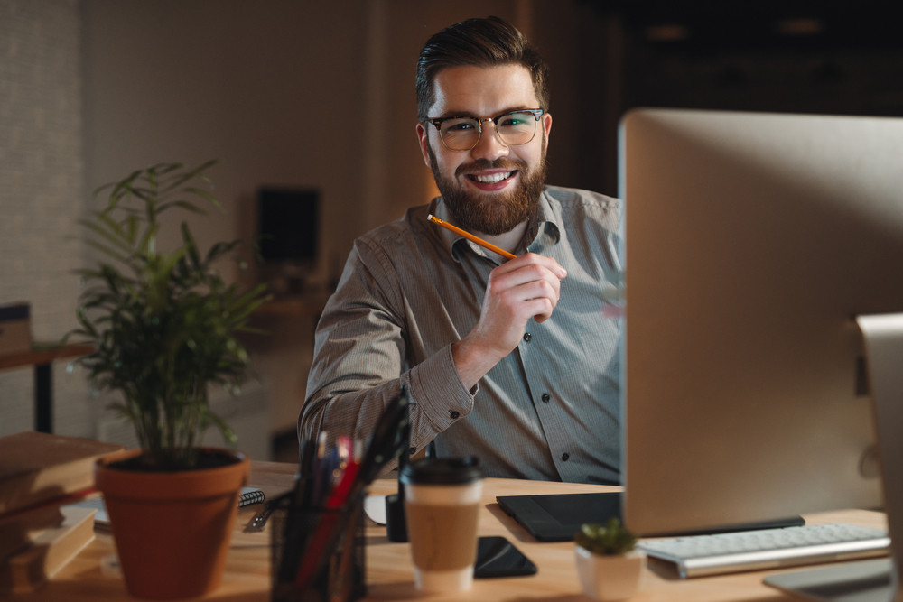Photo of cheerful bearded web designer dressed in shirt working late at night and looking at camera while holding pen in hand.