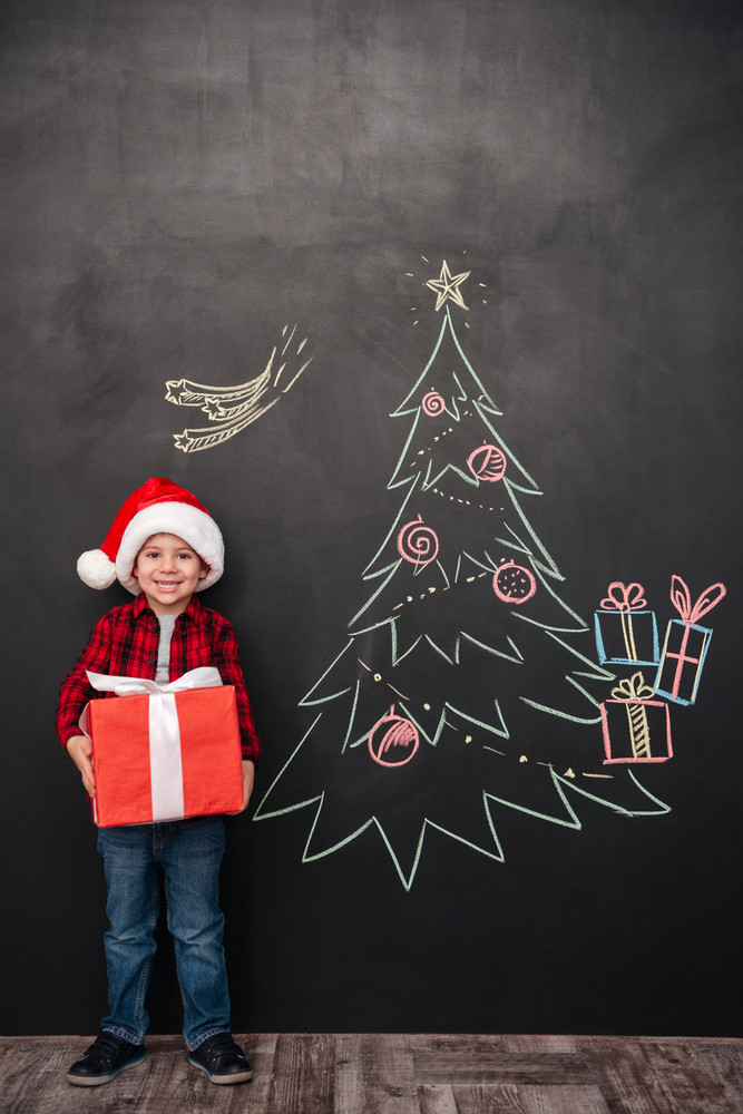 Photo of cheerful child wearing hat holding a big gift near Christmas tree drawing on blackboard. Looking at camera.