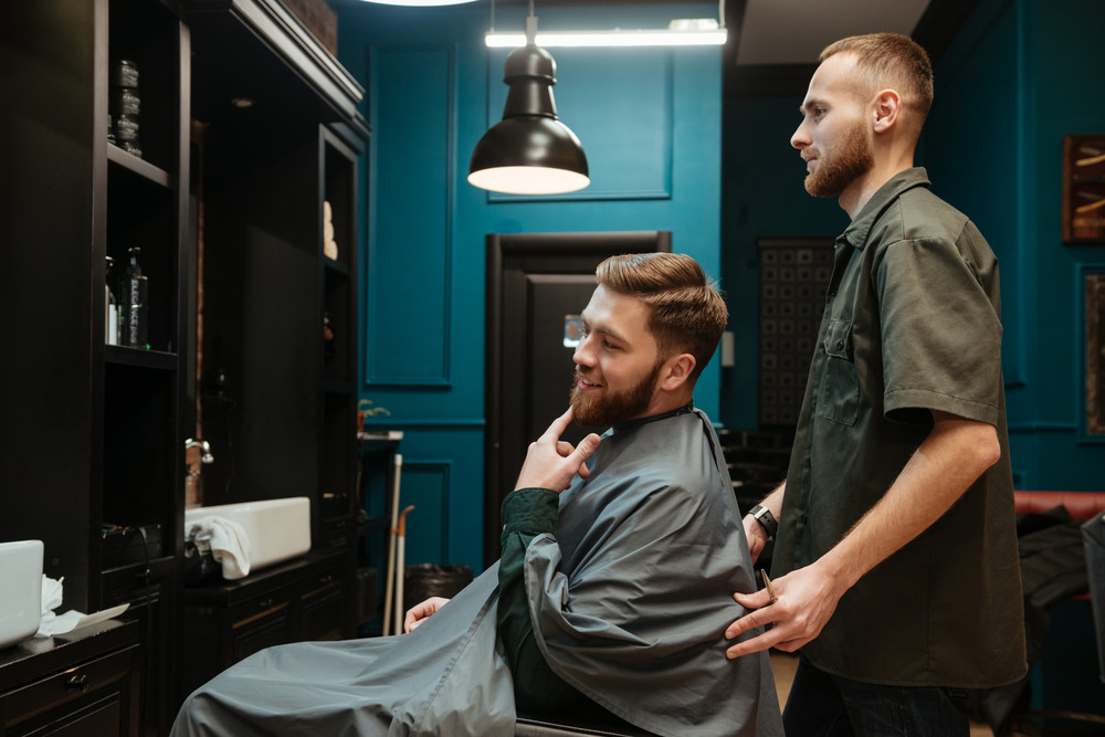Photo of handsome man getting haircut by hairdresser while sitting in chair. Look at mirror.